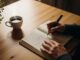 Una persona scrive con una penna su un diario aperto, seduta a un tavolo di legno. Vicino al diario ci sono una tazza di caffè e una pianta in vaso. L'immagine è ripresa dall'alto, con una luce calda che crea un'atmosfera rilassata e riflessiva.
